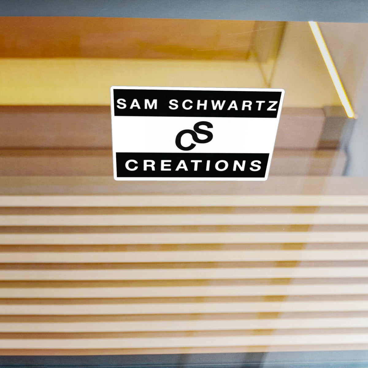 A rectangular black and white sticker with the text 'SAM SCHWARTZ CREATIONS' and a stylized ampersand logo, shown on a glass door with a blurred background of vertical wooden slats.